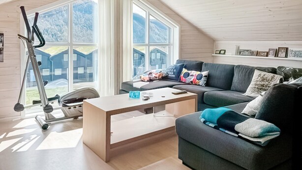 Una sala de estar moderna con un sofá gris, una mesa de centro y una cinta de correr. Amplias ventanas ofrecen vistas a un paisaje montañoso.