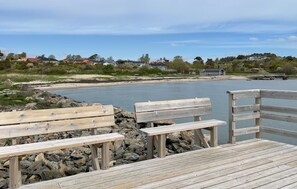 Un muelle de madera con bancos que da a una masa de agua tranquila, con casas y vegetación al fondo.