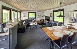 Un interior moderno con techo de madera, un comedor con mesa y sillas de madera, una zona de estar con un sofá curvo y ventanas grandes que ofrecen luz natural.