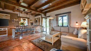 Een ruime woonkamer met een bank, een eethoek met stoelen, een televisie en een keuken die door een open deur zichtbaar is.