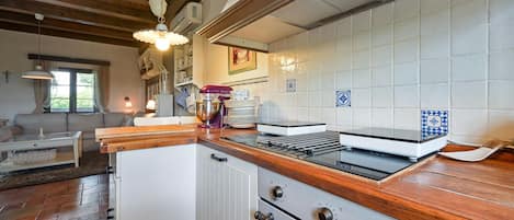 Cozinha com bancada de madeira, armários brancos e forno embutido. Há uma janela com cortinas e um lustre.