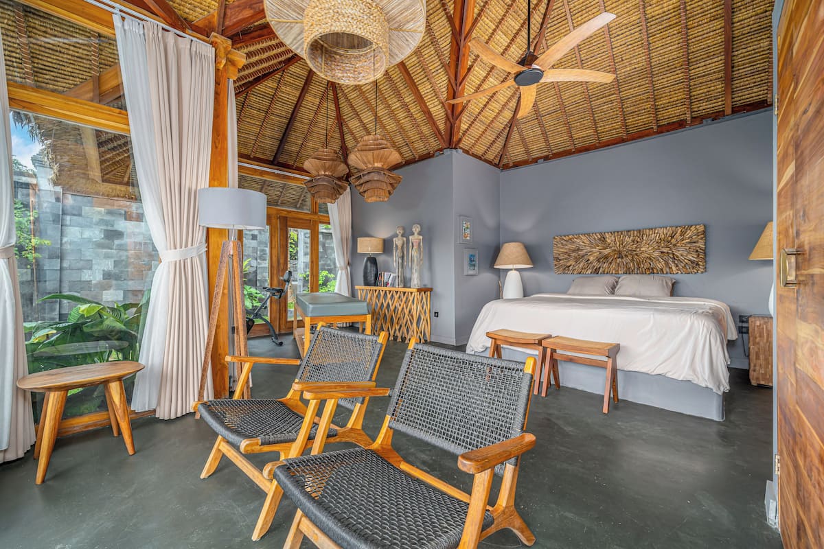 A room with a bed, two chairs, a bench, a table, and a ceiling fan.