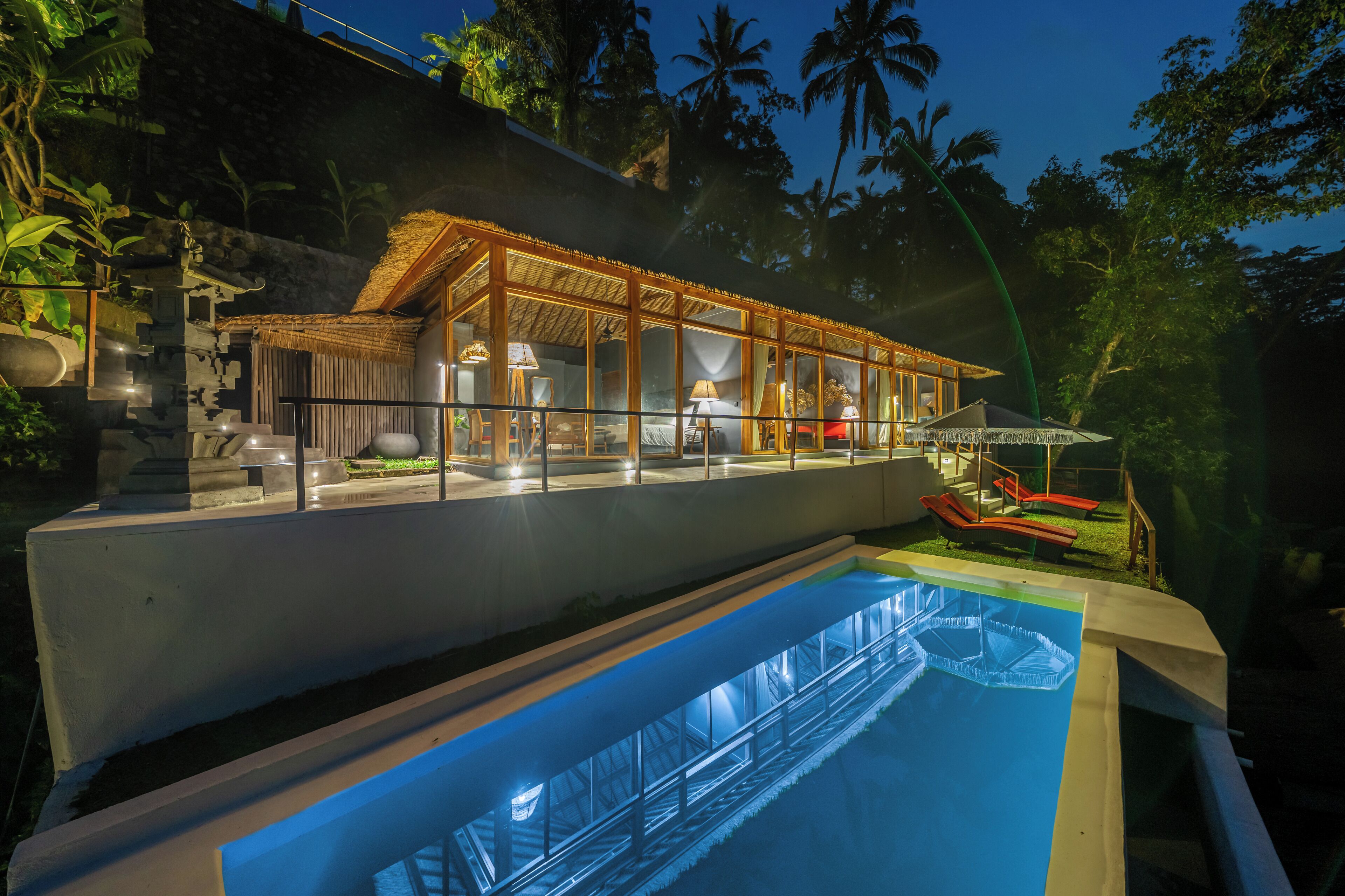 A modern resort with a glass-enclosed pool area, a well-lit lounge, and a view of the surrounding tropical landscape.