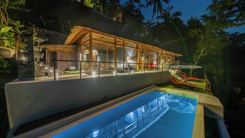 A modern resort with a glass-enclosed pool area, a well-lit lounge, and a view of the surrounding tropical landscape.