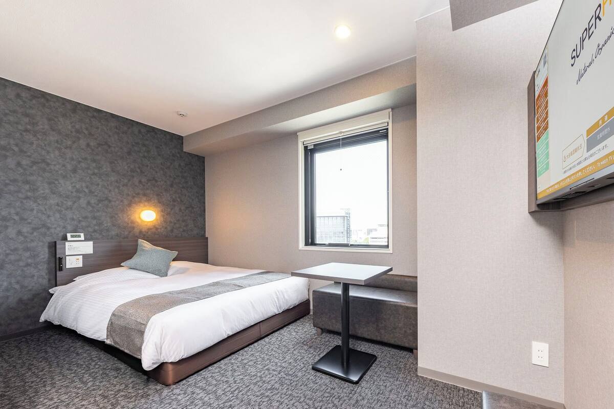 Una habitación de hotel moderna con cama, mesita de noche y un ventanal con vistas a la ciudad.