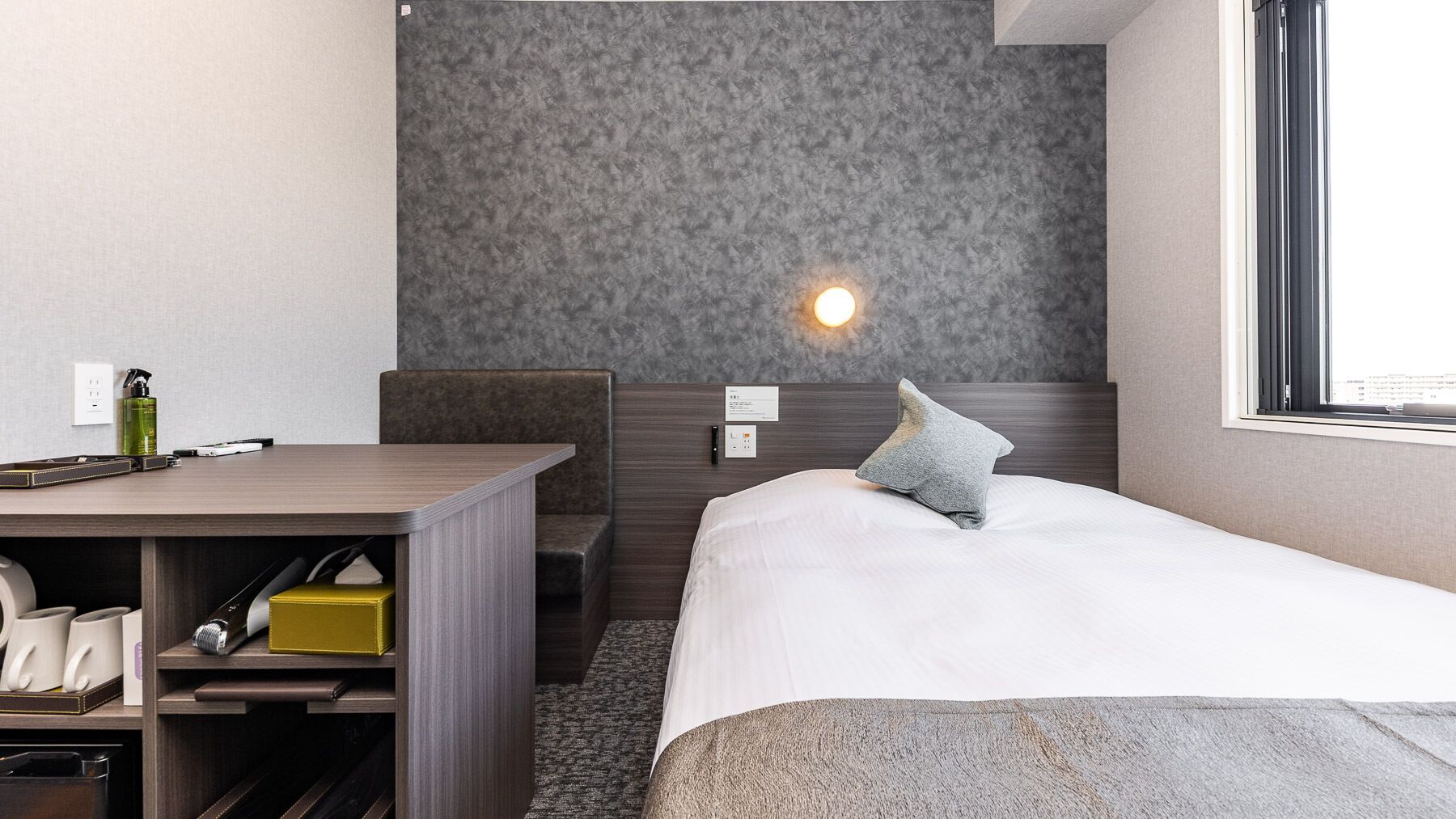 Habitación de hotel con cama, escritorio, silla, mesita de noche, lámpara y ventana.