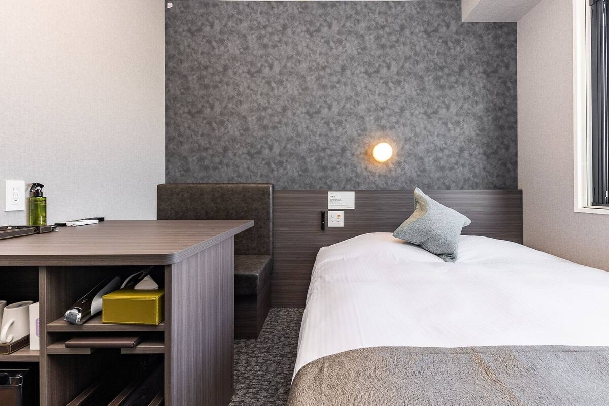 Habitación de hotel con cama, escritorio, silla, mesita de noche, lámpara y ventana.