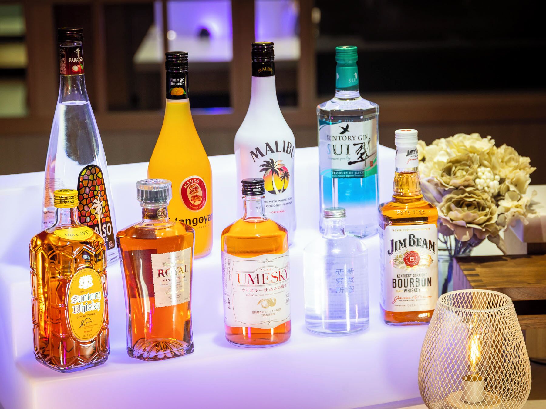 A variety of liquor bottles on a table with a decorative lamp and flowers.