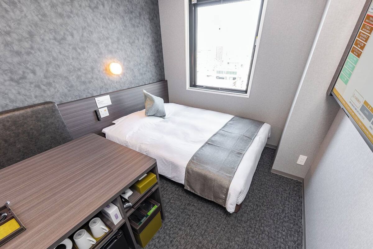 Habitación de hotel compacta con cama, mesita de noche y un pequeño escritorio con espacio de almacenamiento.