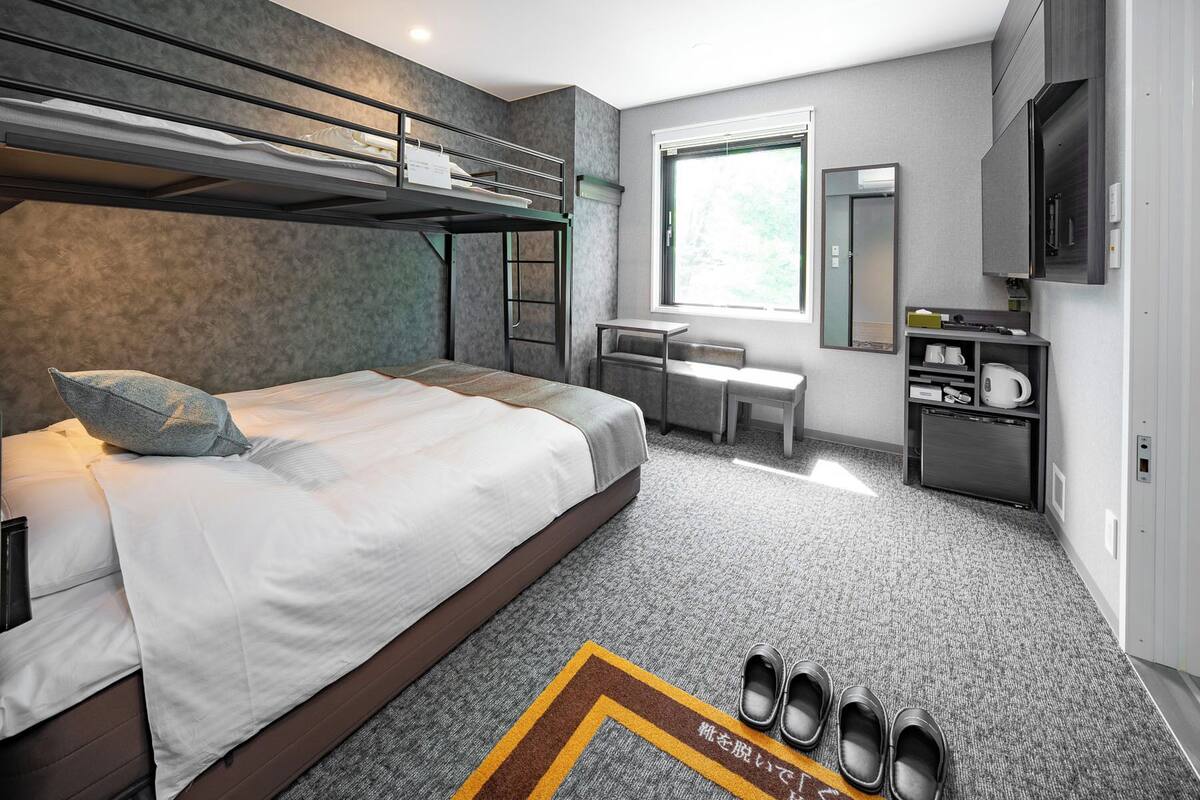 Una habitación de hotel moderna con una cama litera, un escritorio, una silla, una pequeña kitchenette y un ventanal grande.