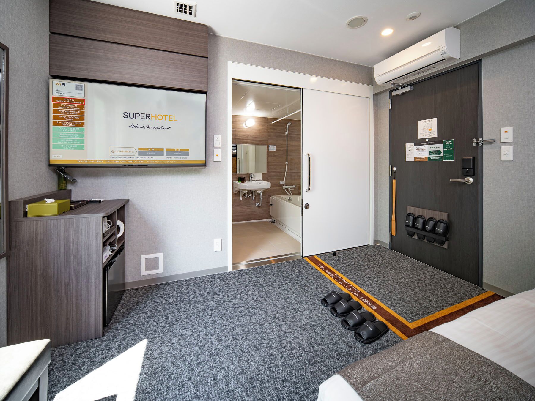 A hotel room with a bed, a desk, a TV displaying room amenities, a bathroom with a shower, and a shoe rack.