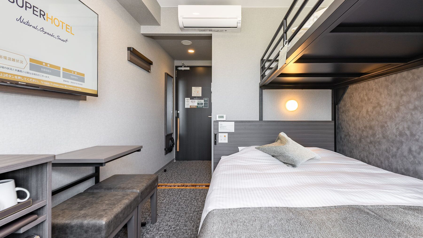 Habitación de hotel con cama, mesita de noche, banco y televisión instalada en la pared.