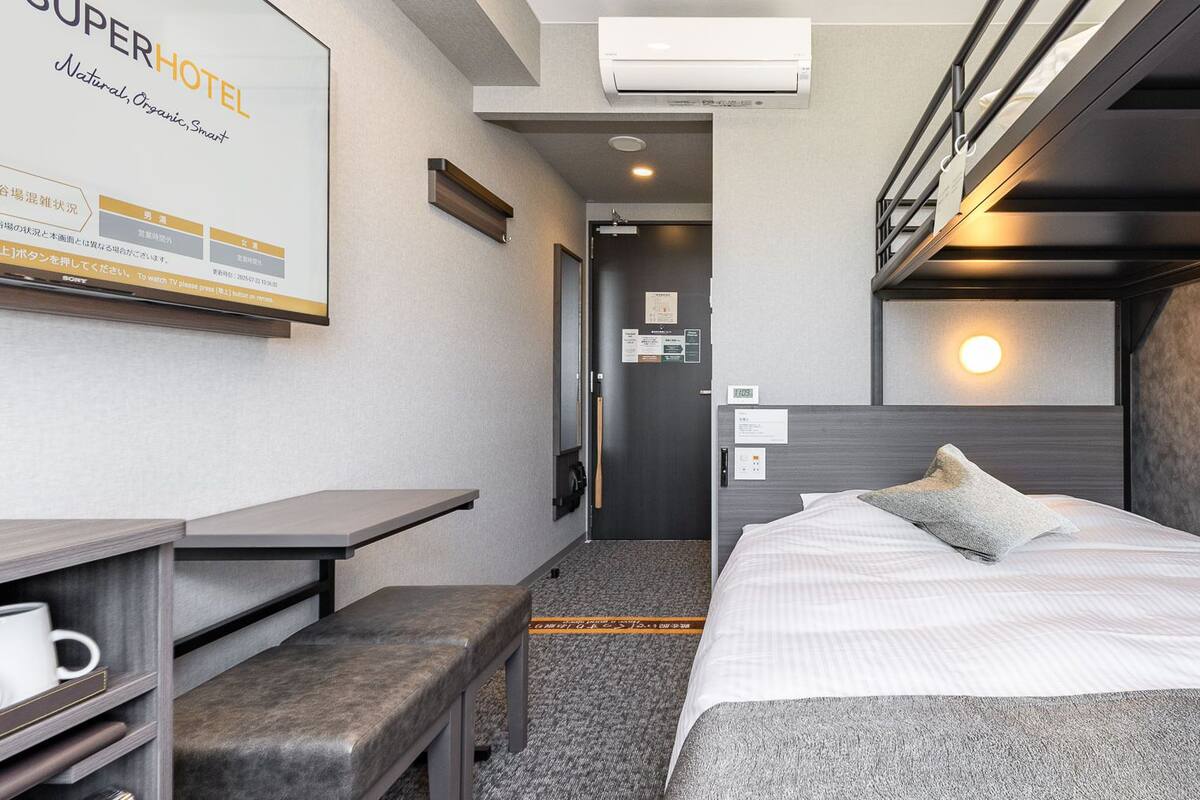 Habitación de hotel con cama, mesita de noche, banco y televisión instalada en la pared.
