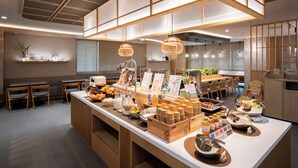 Un restaurante moderno con una barra central que exhibe diversos alimentos y condimentos. Hay mesas y sillas de madera, y el techo presenta un diseño geométrico con iluminación empotrada.