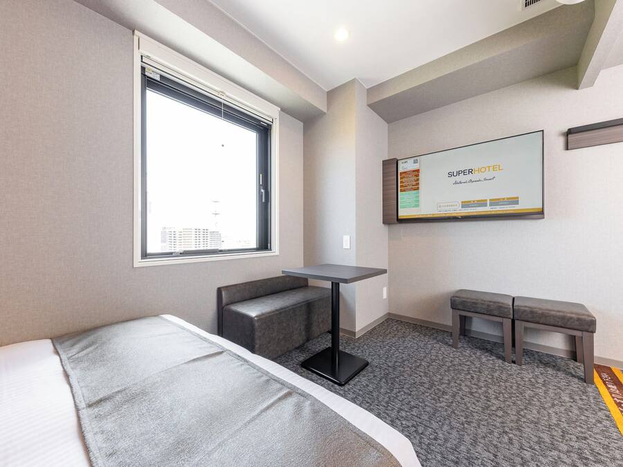 A hotel room with a bed, a small desk, a bench, and a large window.
