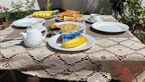 A table set outdoors with a white lace tablecloth, plates with food, a teapot, and a window with white frames. - Silver Meir Bungalow (Nuwara Eliya)