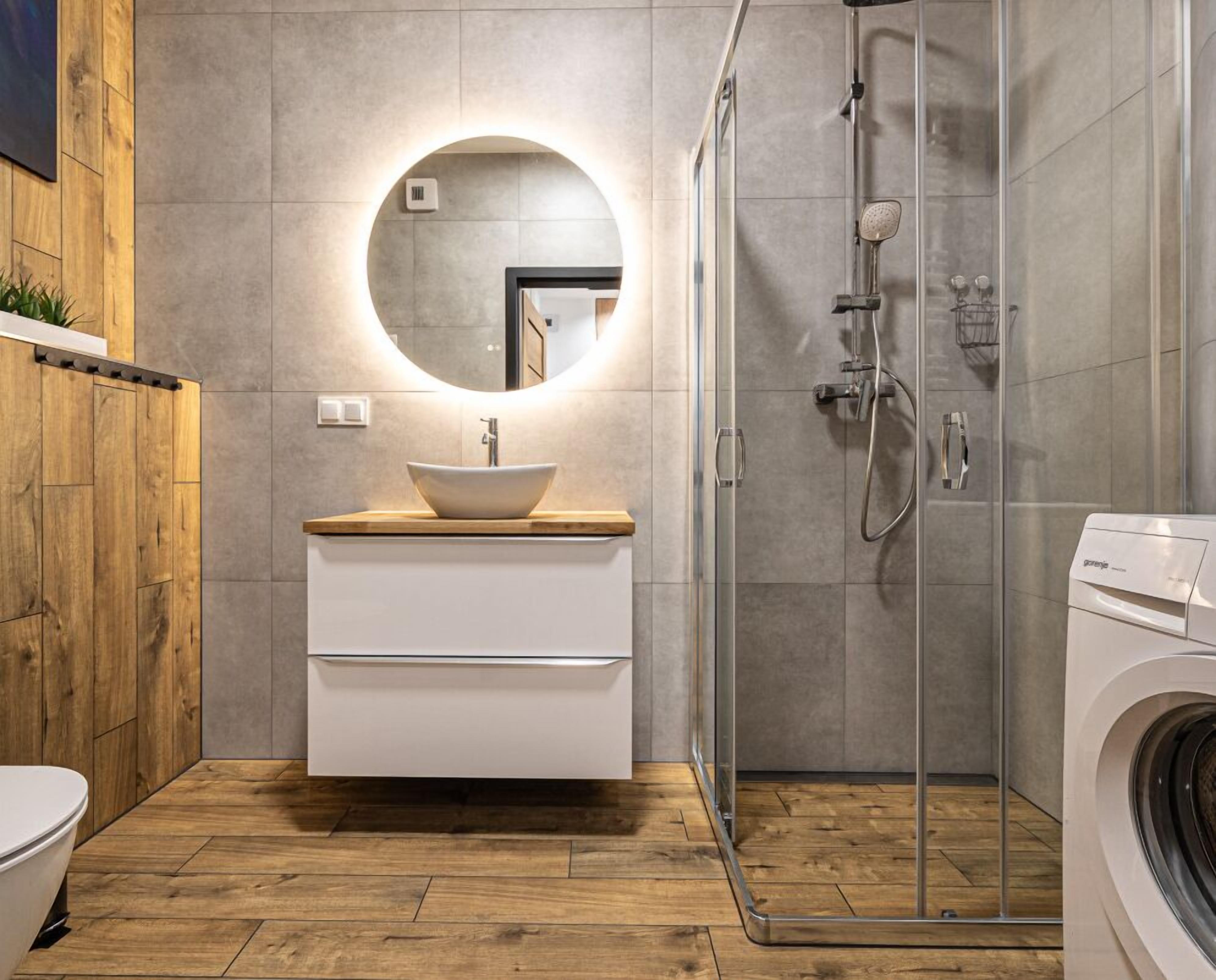 A modern bathroom with a wooden vanity, a round mirror, a glass shower enclosure, and a washing machine.