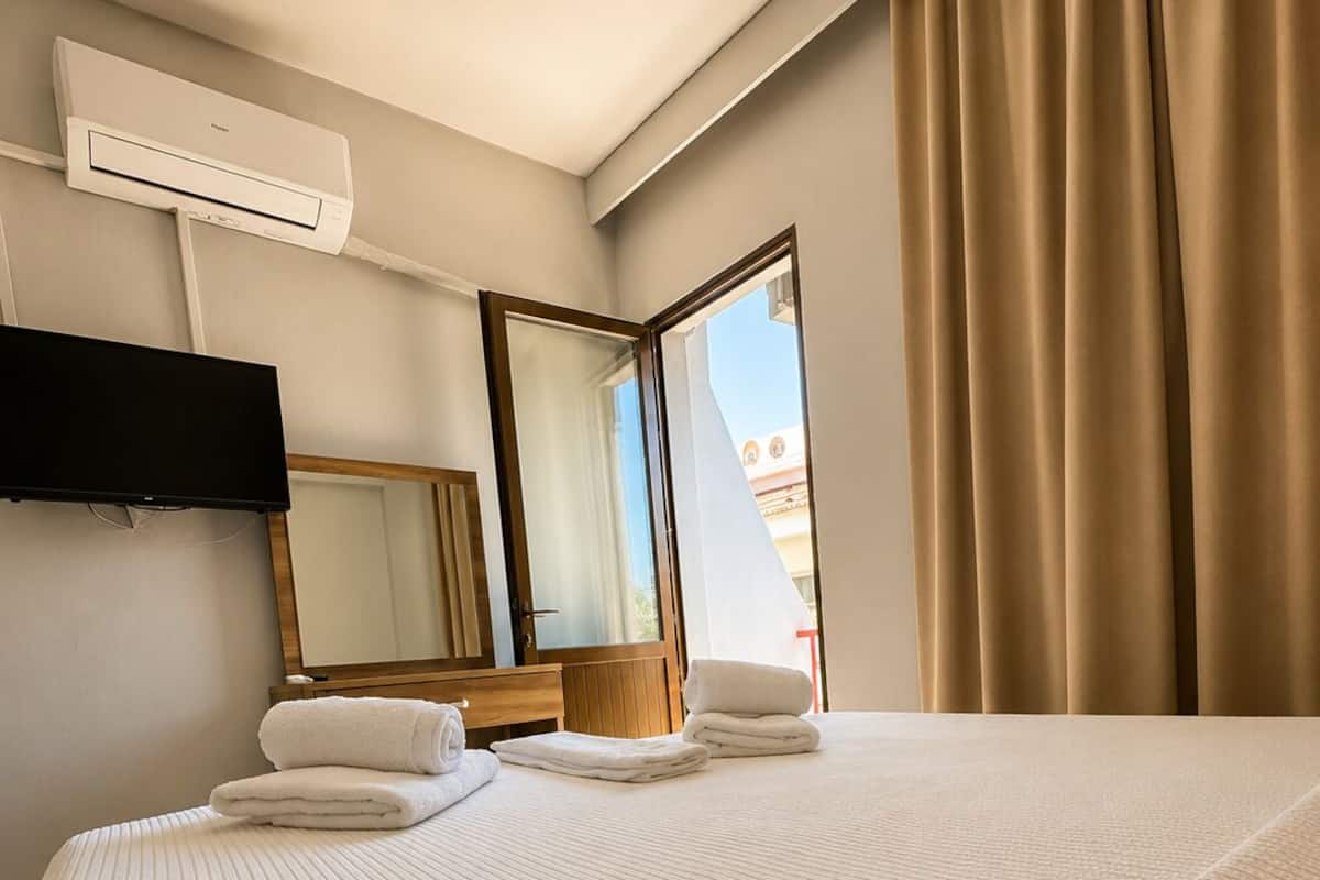 A hotel room with a bed, a flat-screen TV, a mirror, a window with curtains, and an air conditioning unit.