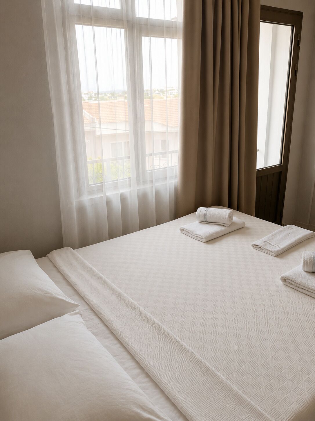 A hotel room with a bed, two pillows, a window with curtains, and a view of buildings outside.