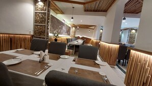 En restaurant med et langt spisebord dækket til fire personer, træpaneler på væggene og udsigt til det frie gennem et vindue.
