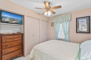 Una camera da letto con un letto, una cassettiera, una ventola a soffitto, una TV su uno scaffale, una finestra con tende e un quadro appeso al muro.