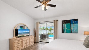 Une chambre avec un lit, un téléviseur posé sur une commode, un ventilateur de plafond et une vue sur une piscine.