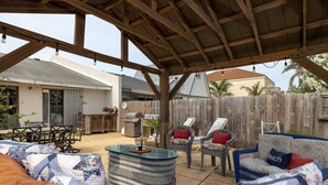Un patio exterior con una pérgola de madera, una mesa con cubierta de vidrio y mobiliario de exterior.