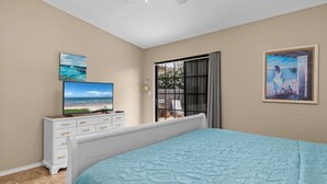 Un dormitorio con cama, televisor sobre una cómoda, ventana con cortinas y un cuadro en la pared.