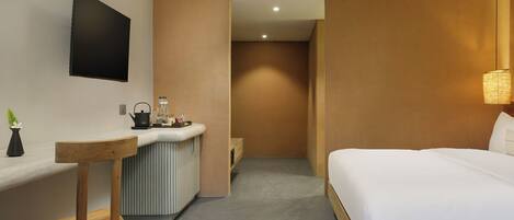 Habitación de hotel con cama, escritorio, silla, kitchenette pequeña y televisor montado en la pared.