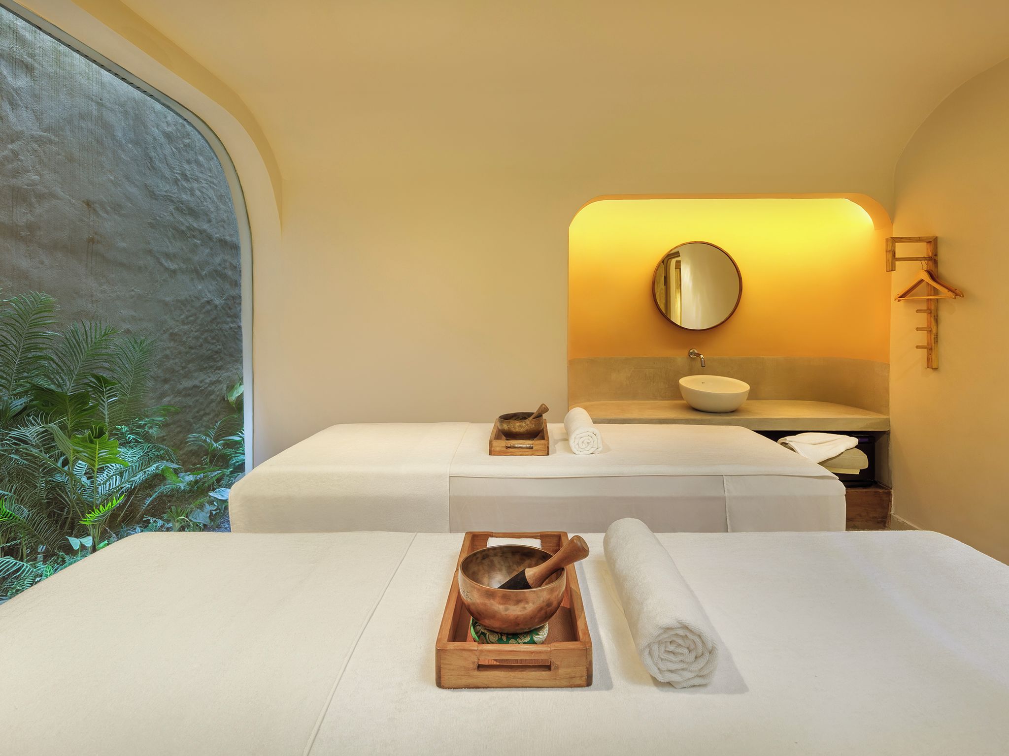 A spa room with two beds, a sink, a mirror, and a wooden tray with a bowl and a rolled towel.