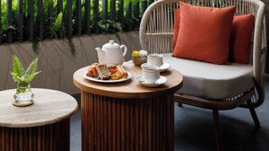 A round wooden table with a white teapot, a cup, and a plate with pastries. A wicker chair with a cushion is in the background. - Bagaincha Resort & Spa (Pokhara)