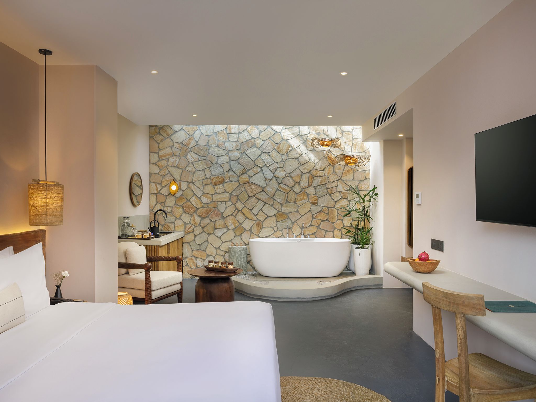 A modern hotel room with a stone wall, a large bed, a sitting area, a bathtub, and a TV mounted on the wall.