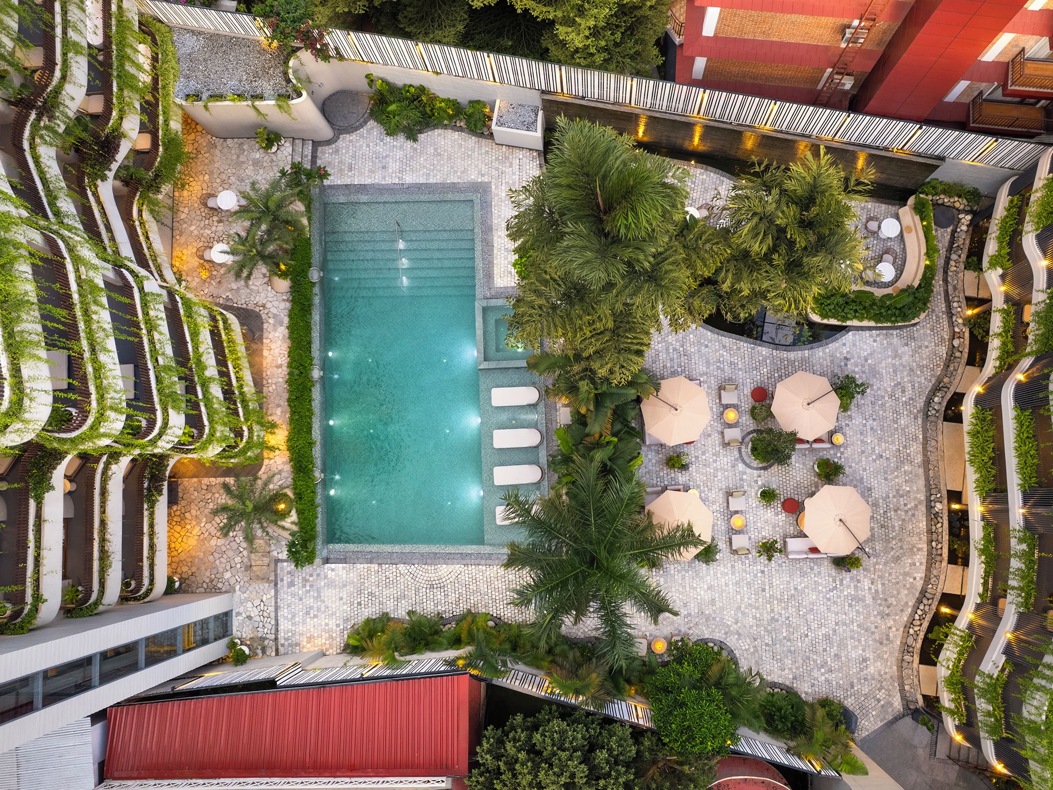 An aerial view of a rooftop pool area with a swimming pool, lounge chairs, and surrounding greenery.