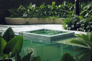 Uma piscina retangular com borda revestida em azulejos, cercada por plantas tropicais exuberantes e um caminho de pedra.
