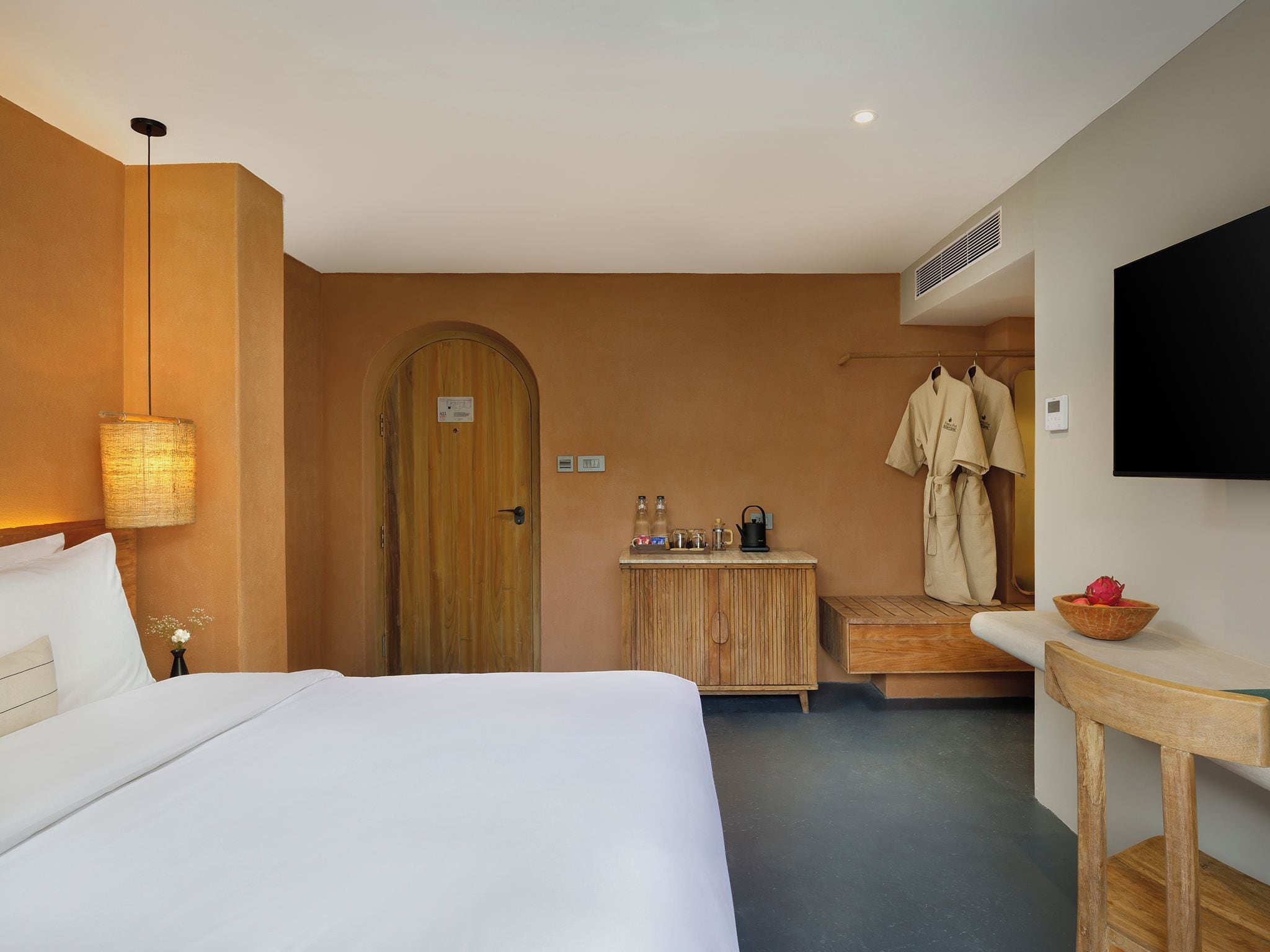 A hotel room with a bed, a wooden door, a small table with toiletries, a robe hanging on the wall, and a television.