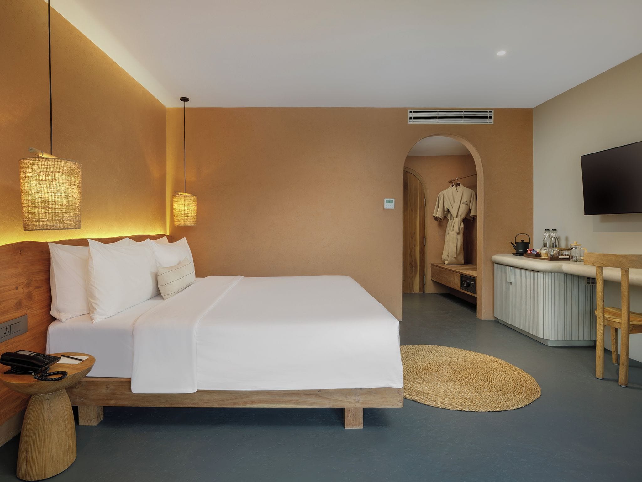 A hotel room with a large bed, bedside lamps, a desk with a flat-screen TV, a robe hanging, and a kitchenette.