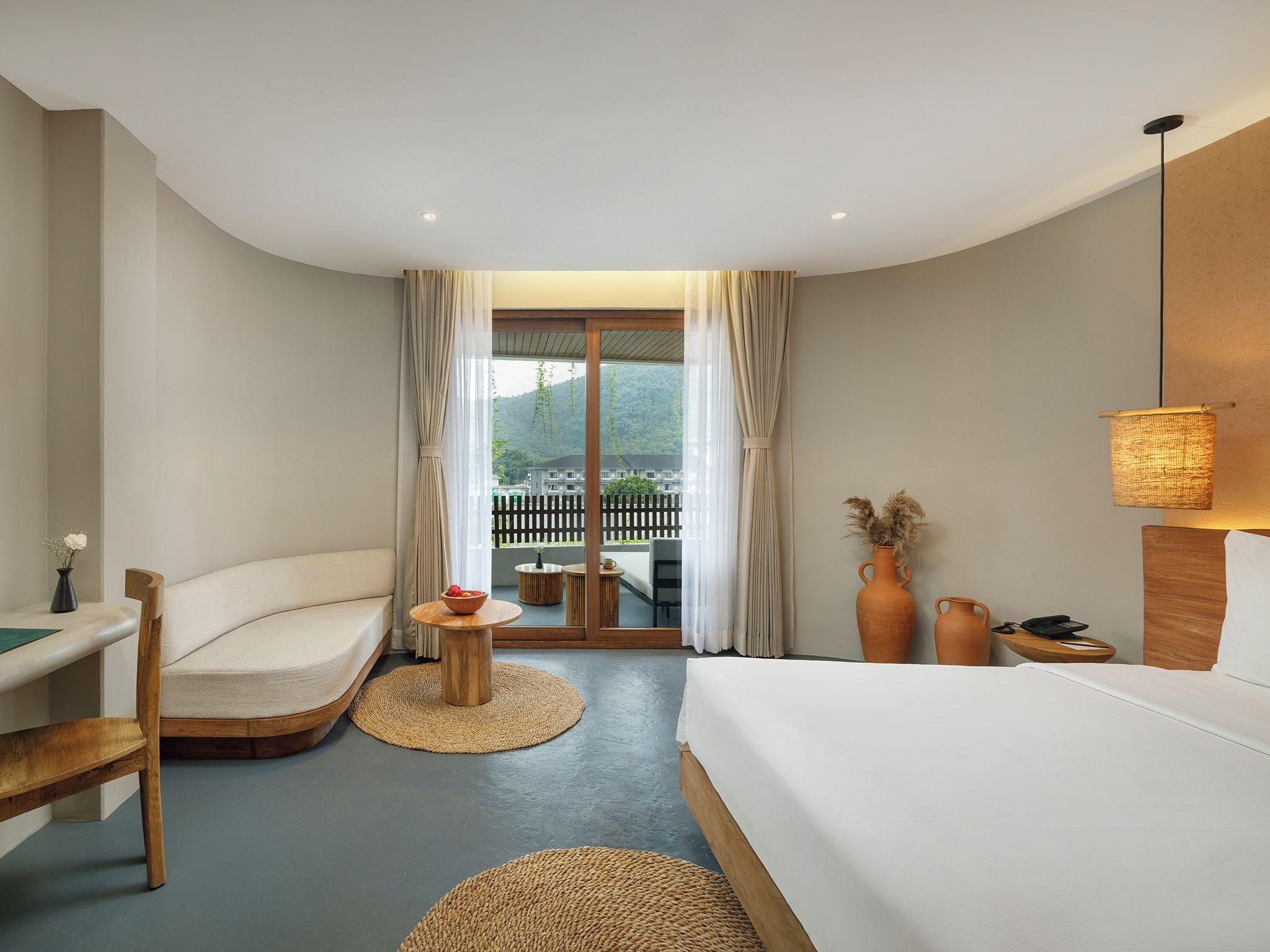 A hotel room with a bed, a sofa, a small round table, a chair, and a view of a balcony with a fence.