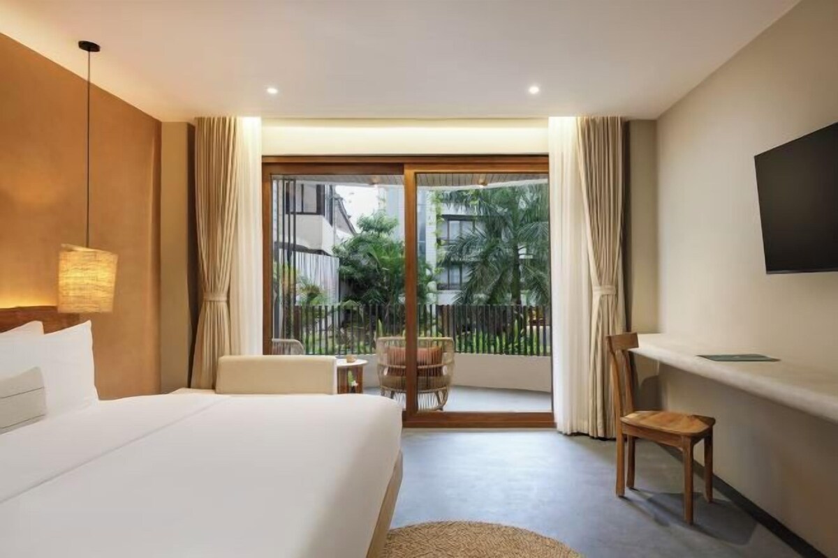 A hotel room with a bed, a desk, a chair, a TV, and a balcony with a view of greenery.