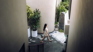 Une femme marchant dans un couloir étroit, bordé de plantes en pot et flanqué d’un mur de pierre.