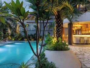 A swimming pool with a stone pathway, surrounded by tropical plants and a wooden bar area.