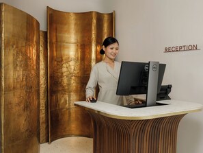 A reception desk with a computer, a woman standing behind it, and a 'RECEPTION' sign on the wall. - Bagaincha Resort & Spa (Pokhara)