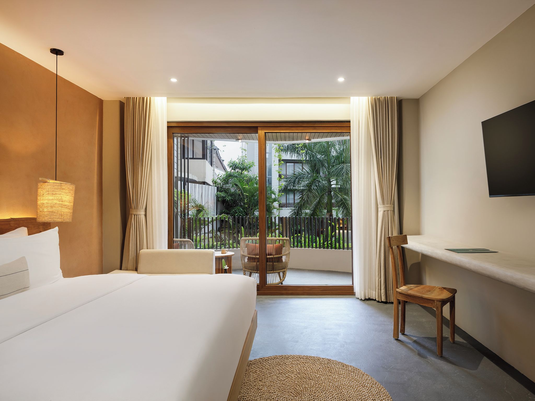 Hotelkamer met een bed, bureau, stoel, televisie en balkon met uitzicht op groen en gebouwen.