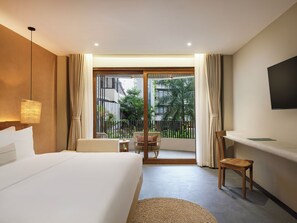 Habitación de hotel con cama, escritorio, silla, televisor y balcón con vistas a zonas verdes y edificios.