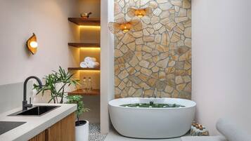 Een moderne badkamer met een vrijstaand bad, een houten wastafelmeubel, een accentmuur van steen en een kleine plant.