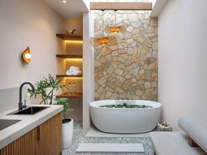 Un baño moderno con una bañera independiente, un tocador de madera, una pared de acento de piedra y una pequeña planta.