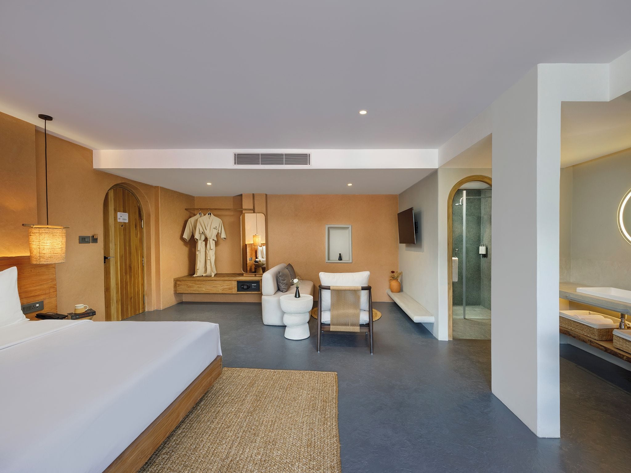 A modern hotel room with a large bed, a seating area with a sofa and ottoman, a small table with a vase, a television mounted on the wall, and a separate bathroom with a glass shower enclosure.