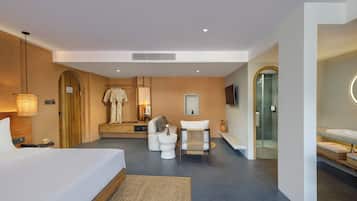 A modern hotel room with a large bed, a seating area with a sofa and ottoman, a small table with a vase, a television mounted on the wall, and a separate bathroom with a glass shower enclosure.