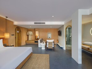 A modern hotel room with a large bed, a seating area with a sofa and ottoman, a small table with a vase, a television mounted on the wall, and a separate bathroom with a glass shower enclosure. - Bagaincha Resort & Spa (Pokhara)