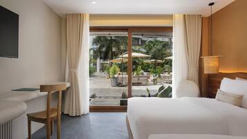 A hotel room with a bed, a desk, a chair, and a view of an outdoor area with plants and umbrellas.