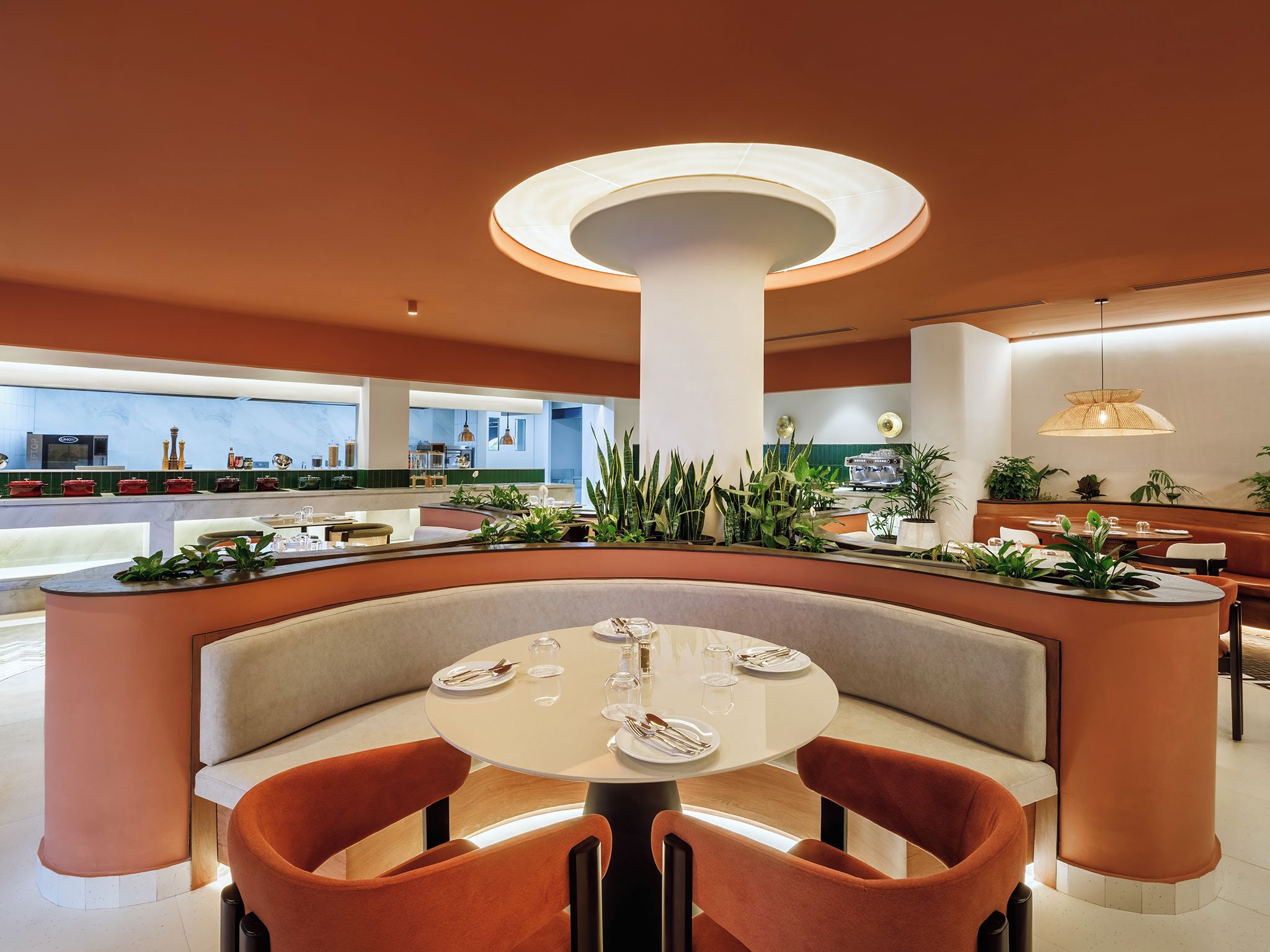 A modern restaurant interior with a central round table, cushioned seating, and a variety of plants.